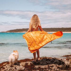 Een grote Sea to Summit Drylite Towel reishanddoek in de kleur Outback Sunset wappert op het strand naast een hond.