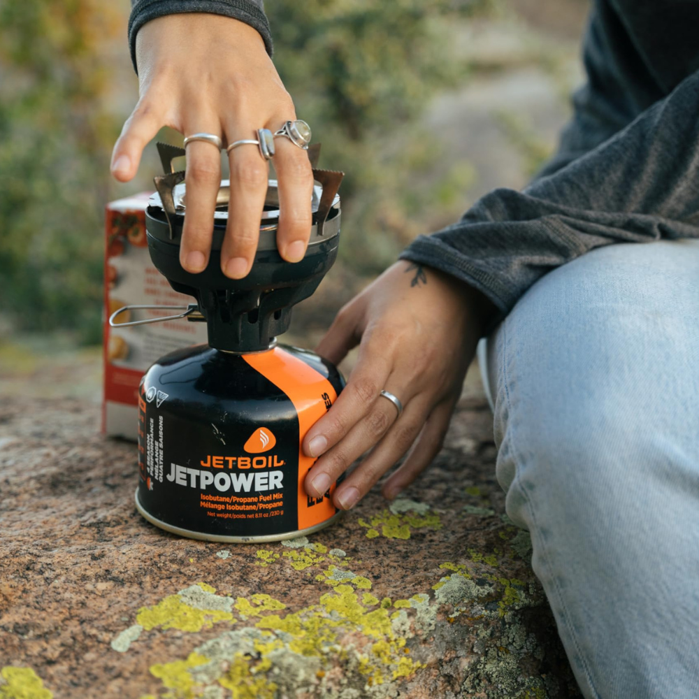 Iemand schroeft een compacte Jetboil outdoorbrander op een Jetpower gasbusje dat op een rots staat.