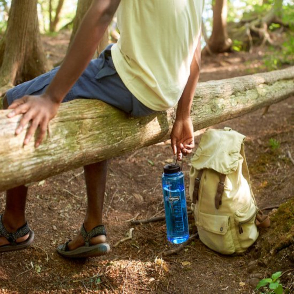 Persoon met een blauwe Nalgene kunststof drinkfles en rugzak zittend op een boomstam in het bos