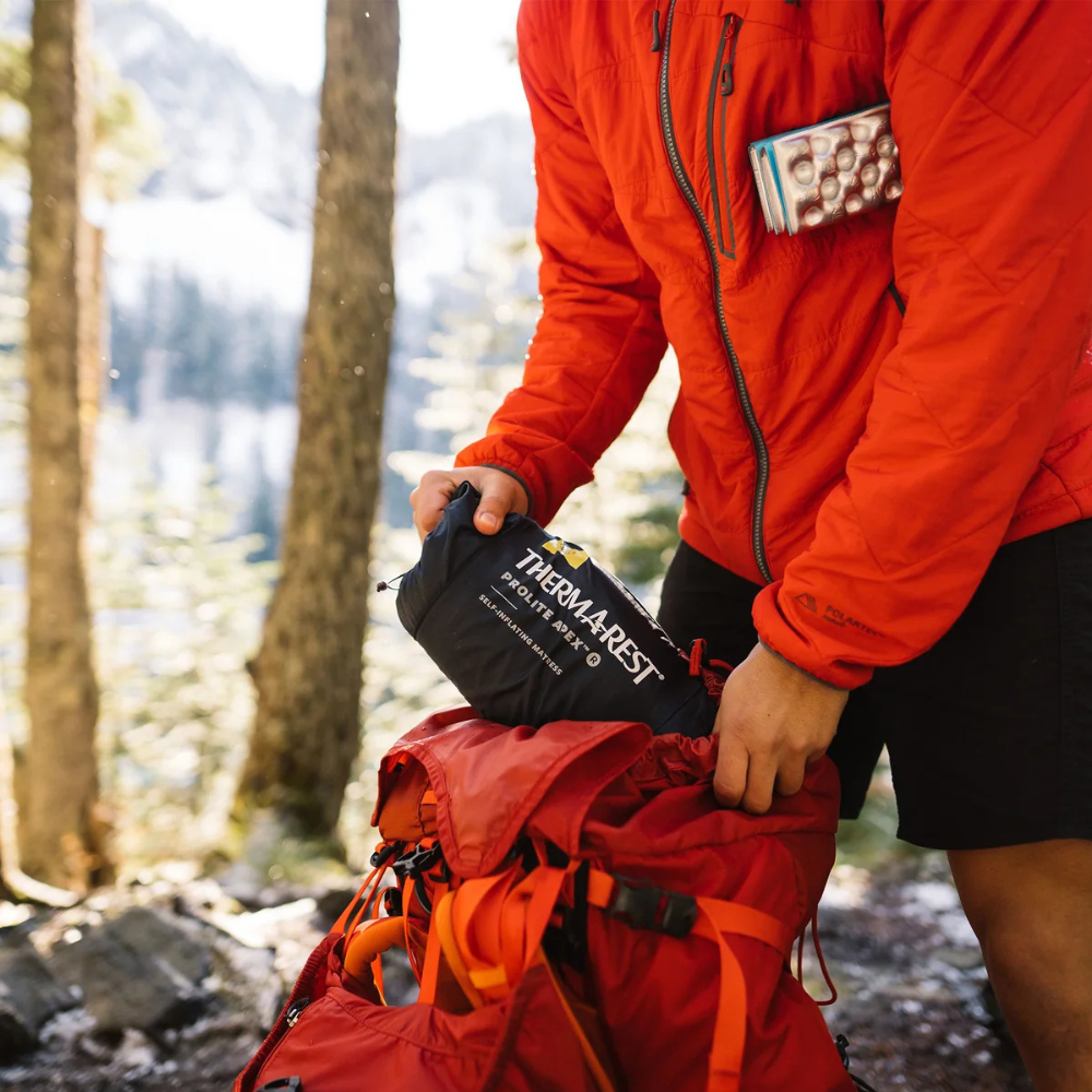  Persoon stopt de opgerolde Therm-a-Rest ProLite Apex Heat Wave L slaapmat in een rode rugzak   
